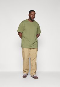 Olijfgroene T-shirt met korte mouwen en ronde hals, gecombineerd met tan cargobroek. Donkere schoenen met witte accenten op een effen achtergrond.