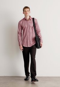 Camicia a quadri rossi e bianchi, borsa a tracolla nera, pantaloni scuri, scarpe nere, in piedi su un pavimento chiaro contro un muro semplice.