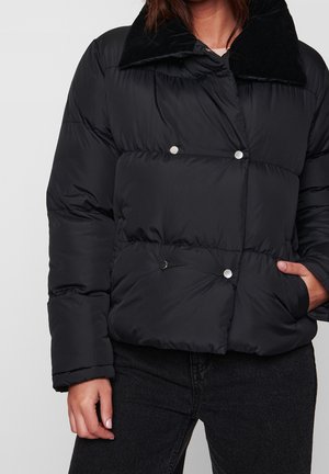 Femme portant une veste matelassée noire avec des boutons-pression argentés, un col noir, et un jean noir, une main dans la poche, devant un fond uni.