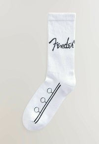 Soseta albă din bumbac cu textură canelată, având un logo negru "Fender" și trei modele circulare deasupra unei benzi orizontale negre.
