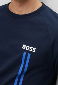 Uomo che indossa una camicia blu marina con il testo bianco "BOSS" e due strisce verticali blu sotto il testo sul petto.