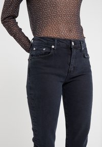 Jean en denim noir avec une coupe ajustée, cinq poches, une couture apparente et des accents de quincaillerie argentée. Le matériau semble texturé et légèrement usé.