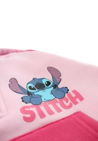 Sweat-shirt rose avec un graphique cartoon de Stitch en bleu, assis les mains levées, et le mot "STITCH" en texte rose en gras en dessous.
