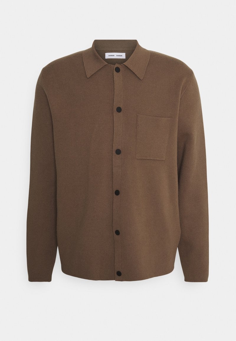 Chemise marron à manches longues avec boutons noirs et une seule poche poitrine, présentée sur un fond blanc uni.