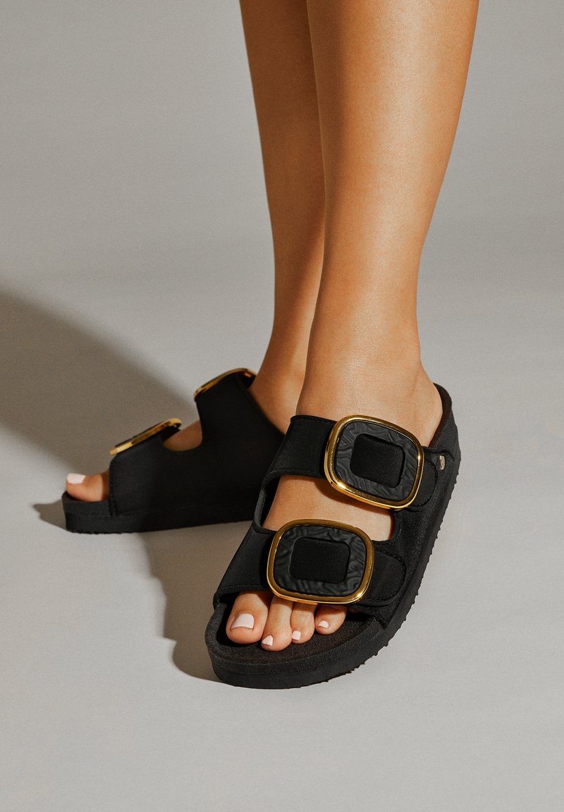 Chanclas negras con dos grandes hebillas cuadradas en oro y acentos texturizados en negro, material suave y suela plana de goma.