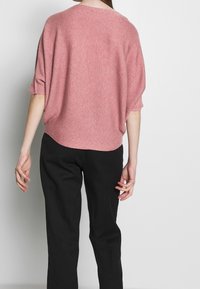 Rosa Strickpullover mit lockerer Passform, überschnittenen Schultern und kurzen Ärmeln; der Stoff hat eine gerippte Textur; kombiniert mit schwarzen Hosen.