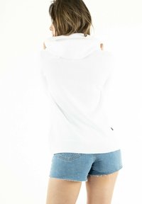 Donna con capelli castani che indossa un hoodie bianco e pantaloni di jeans blu, girata di spalle, mentre tiene il cappuccio con entrambe le mani su uno sfondo bianco.