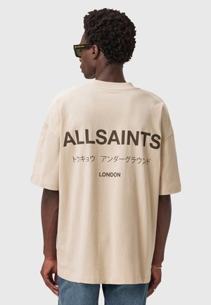 Beige oversized t-shirt with dark text reading "ALLSAINTS," Japanese characters, and "LONDON" on the back; cotton material with a smooth texture.