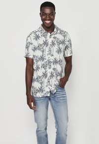 Camicia a maniche corte con bottoni di colore bianco, decorata con una stampa di foglie di palma nere. Indossata con jeans azzurri chiari, dal taglio rilassato e con lievi scoloriture.