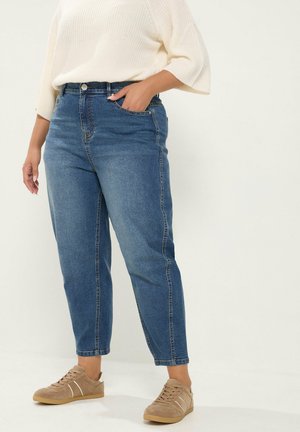Person trägt blaue High-Waist-Jeans, ein weißes gestricktes Top und beige Sneaker und steht vor einem schlichten hellen Hintergrund.