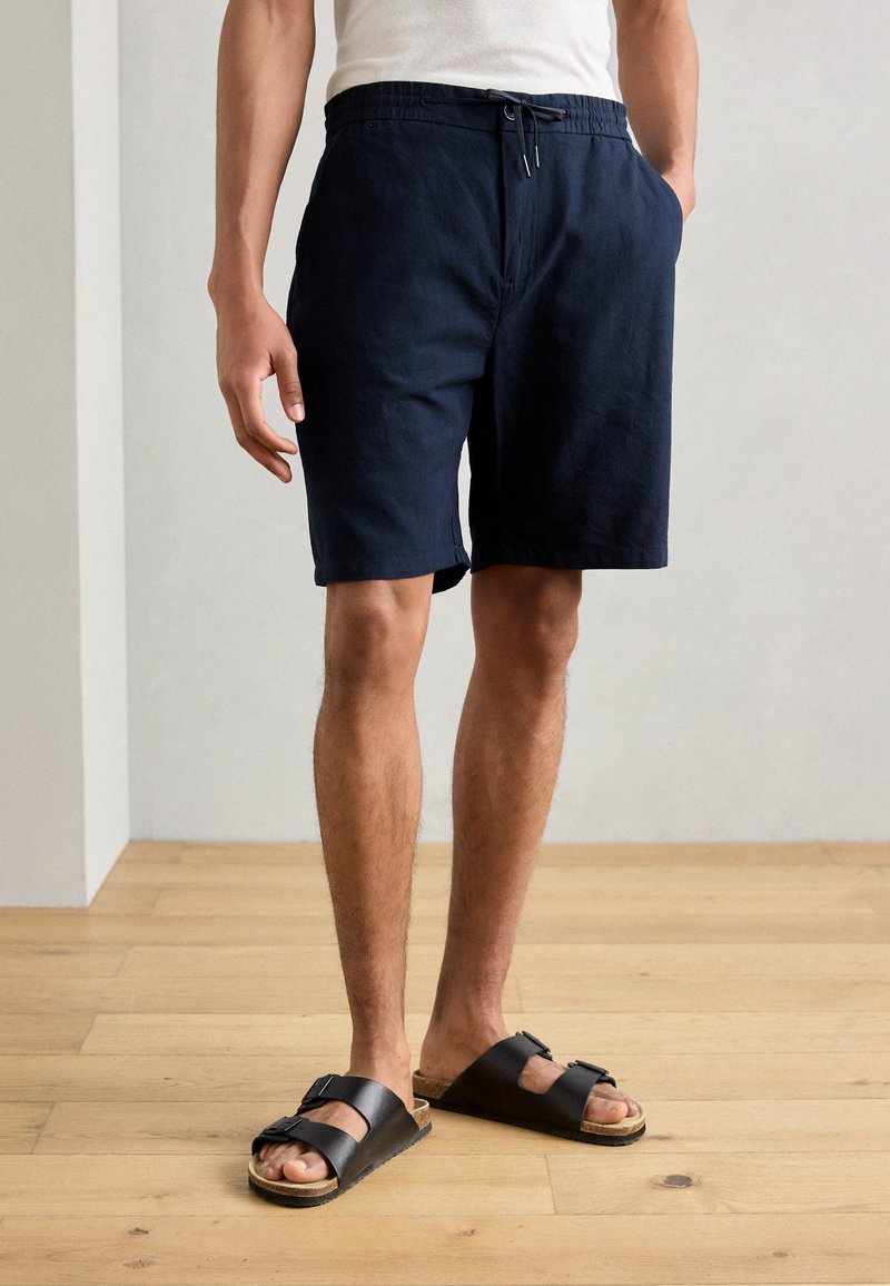 Pantaloni scurți din in de culoare bleu marin, cu talie elastică și șnur, având buzunare laterale și o croială lejeră; asortați cu sandale negre.