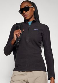Black fleece pullover with a textured pattern, half-zip closure, and a small logo on the chest, paired with khaki pants and carrying a backpack.