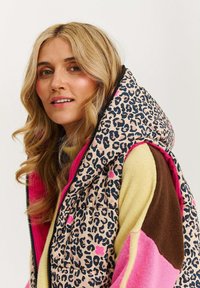 Vesta s leopardím potiskem, polstrovaná, s růžovým vnitřním podšívkou a kapucí. Obsahuje černý zip a svetr v několika barvách s žlutými, hnědými a růžovými částmi.