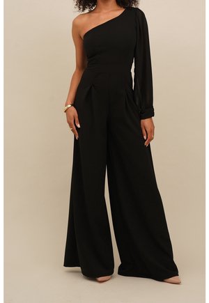 Kvinde iført sort asymmetrisk jumpsuit med vide ben og lang ærme på venstre arm, står mod en beige baggrund.