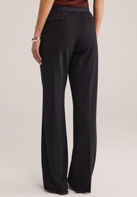 Femeie purtând pantaloni negri largi cu talie înaltă și buzunare cu fante pe spate, alături de un top maro, stând în fața unui fundal simplu.