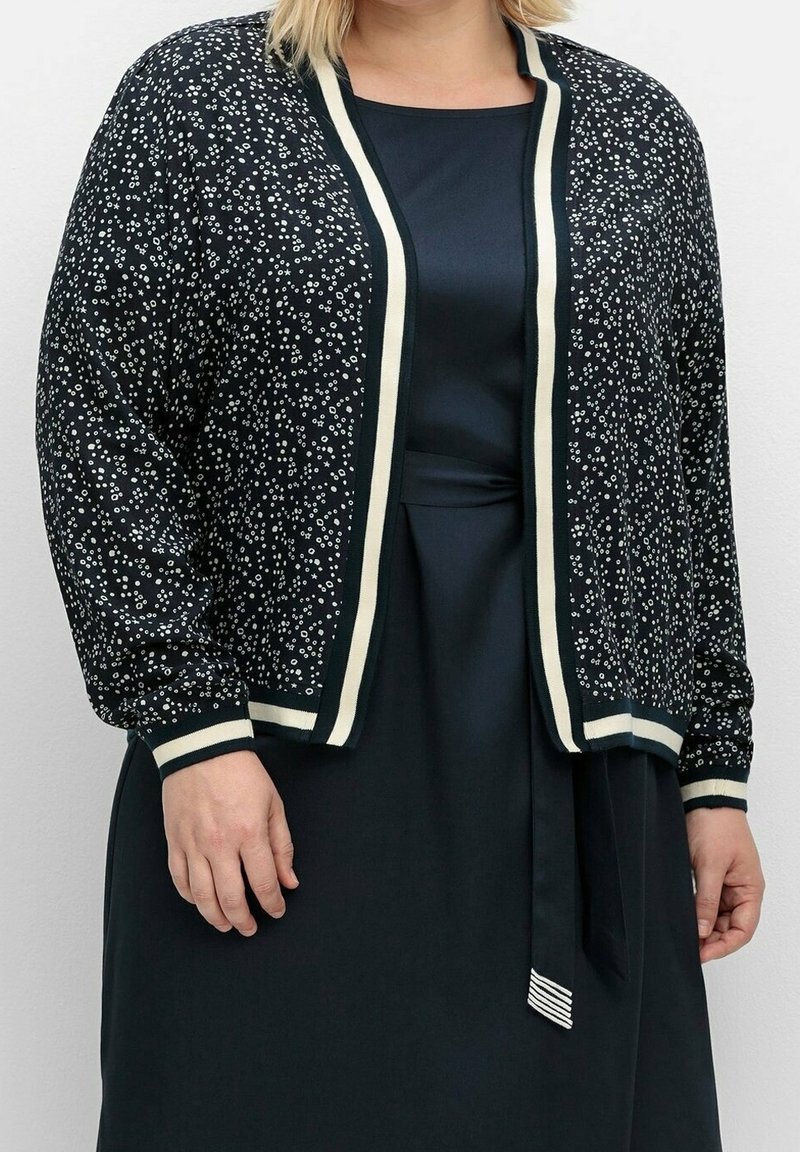 Cardigan a righe bianco e nero con maniche lunghe, polsini a coste e bordo crema lungo i bordi; indossato sopra un vestito scuro.
