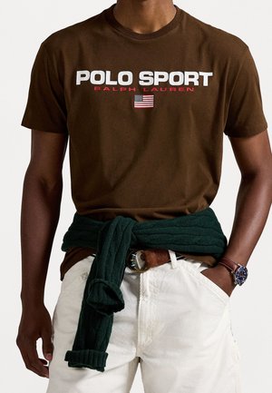 Homme portant un T-shirt marron "Polo Sport Ralph Lauren" avec le drapeau américain, un pantalon blanc, un pull vert noué autour de la taille et une montre au poignet.