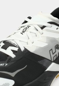 Κοντινό πλάνο σε ασπρόμαυρο παπούτσι τρεξίματος Hoka με λευκά κορδόνια και εμφανή λογότυπα της μάρκας στη γλώσσα και στο πλάι.