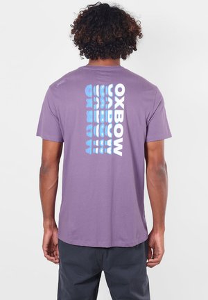 Homme aux cheveux bouclés portant un t-shirt violet avec un texte vertical bleu et blanc indiquant « "OXBOW" » dans le dos, debout devant un fond uni.
