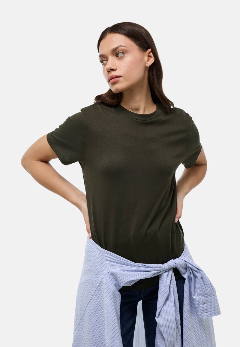 T-shirt olive à manches courtes en tissu lisse, assorti à une chemise rayée bleu clair nouée à la taille, et un jean foncé.