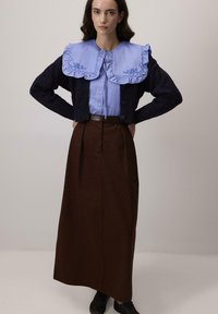 Femeie în picioare, cu mâinile pe șold, purtând o bluză albastră cu guler mare brodat, cardigan negru, fustă lungă maro și pantofi negri.