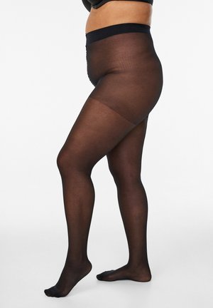 Kobieta ubrana w czarne, przeźroczyste rajstopy stoi bokiem, z jedną stopą lekko uniesioną na jednolitym białym tle.