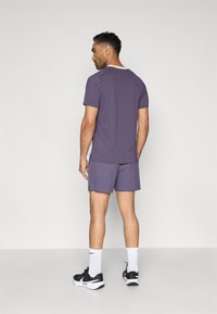 Άνδρας που βλέπει μακριά, φορώντας ένα μοβ κοντομάνικο πουκάμισο, αντίστοιχα καρό σορτς, λευκές κάλτσες Nike και μαύρα παπούτσια Nike.