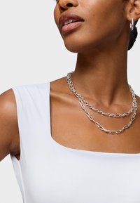 Collana di catena argentata con più fili intrecciati, indossata sopra una maglietta bianca liscia con scollatura quadrata. La tonalità della pelle è visibile.