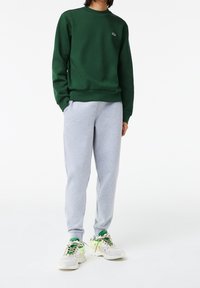 Persona vistiendo un sudadera verde oscuro, pantalones de chándal gris claro y zapatillas blancas con acentos verdes y amarillos, de pie frente a un fondo blanco.