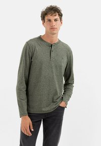 Langærmet henley-skjorte i mørkegrønt stof med en tekstureret overflade, der har en tre-knaps polo og tætsiddende manchetter. Båret med mørke jeans.