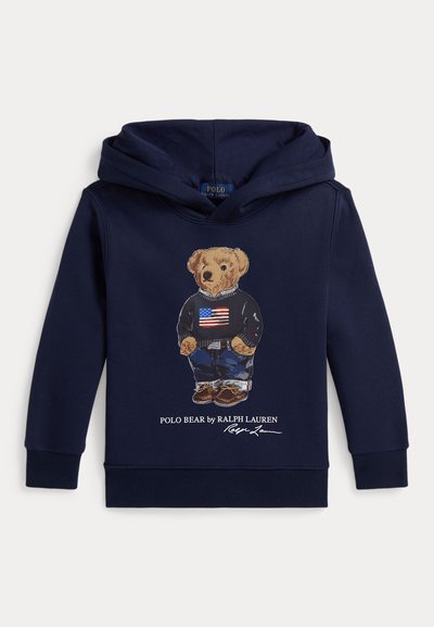 Felpa blu navy con cappuccio con un orso che indossa un maglione nero con la bandiera americana, jeans e scarpe marroni, etichettata "Polo Bear by Ralph Lauren."