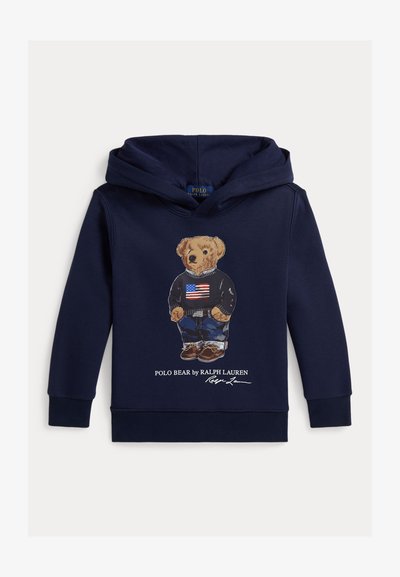 Felpa blu navy con cappuccio con un orso che indossa un maglione nero con la bandiera americana, jeans e scarpe marroni, etichettata "Polo Bear by Ralph Lauren."