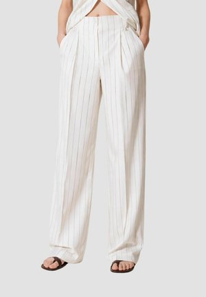 Pantaloni bianchi a vita alta a gamba larga con sottili strisce verticali nere, abbinati a sandali neri con anello per l'alluce, mani nelle tasche.