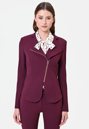Femme portant un blazer prune cintré avec une fermeture éclair asymétrique et un chemisier blanc à pois avec un nœud papillon, debout devant un fond uni.