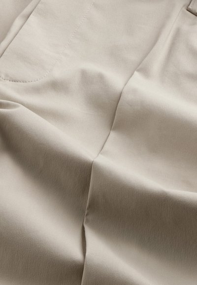 Beige tyg med en jämn textur, med synliga sömmar och subtila veck. Representerar ett plagg, troligen byxor eller shorts.