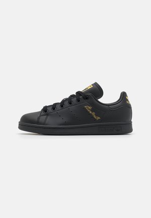 Czarny sneaker Adidas Stan Smith ze złotym logo i charakterystycznymi akcentami, niska konstrukcja i czarne sznurowadła na białym tle.
