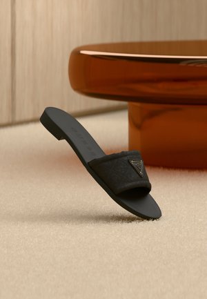 Μαύρο σανδάλι με φιάπα με ραφές και τριγωνικό λογότυπο, τοποθετημένο όρθιο σε μπεζ χαλί κοντά σε στρογγυλό καφέ τραπέζι.