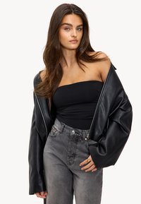 Veste oversize en cuir noir avec des manches larges, portée sur un haut noir ajusté et un jean gris taille haute avec fermeture éclair à l'avant et poches.