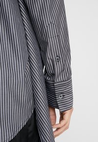 Grå långärmad skjorta med vita vertikala ränder, med knappade manschetter, kontrasterande tyg vid handleden och en böjd fåll.