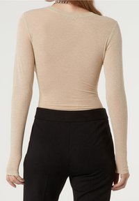 Beige långärmad topp i genomskinlig, texturerad tyg. Har en rund halsringning och en figurnära silhuett, kombinerad med höga svarta byxor.