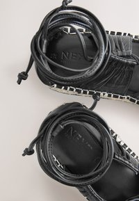 Sorte lædersandaler med fletdesignet, der har sorte snørebånd der er snoet ovenpå og hvide syninger langs sålen. Overfladen har en tekstureret detalje.