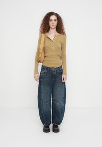 Μπλούζα henley με μακριά μανίκια σε μουσταρδί χρώμα με κουμπιά, συνδυασμένη με υπερμεγέθη μπλε τζιν και μαύρα chunky παπούτσια, κρατώντας μια μπεζ τσάντα.