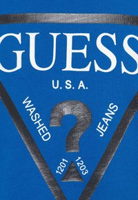 Tela azul con el logo blanco "GUESS" en negrita, un triángulo negro contrastante, "WASHED JEANS" y un diseño de signo de interrogación. Superficie texturizada.