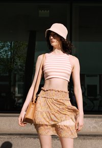 Top cropped riscado em rosa claro e branco, combinado com uma saia mini floral em camadas. Uma bolsa castanha clara e um chapéu de balde bege completam o visual.