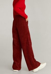 Pantalons larges en velours côtelé rouge avec taille haute et poches latérales, portés avec des baskets blanches. Le tissu présente des côtes verticales et une texture douce.