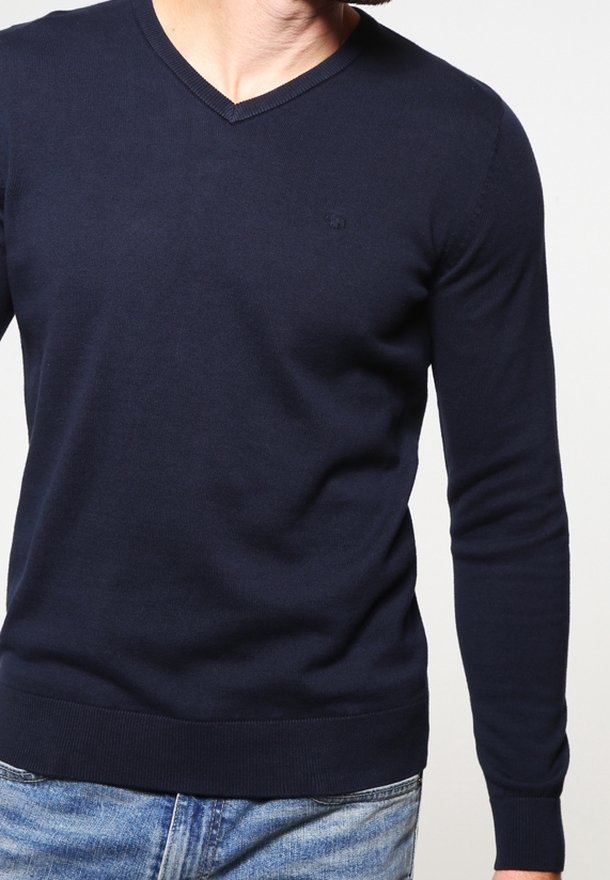 Homme portant un pull bleu marine ajusté col en V à manches longues et un jean bleu clair, debout devant un fond uni et clair.