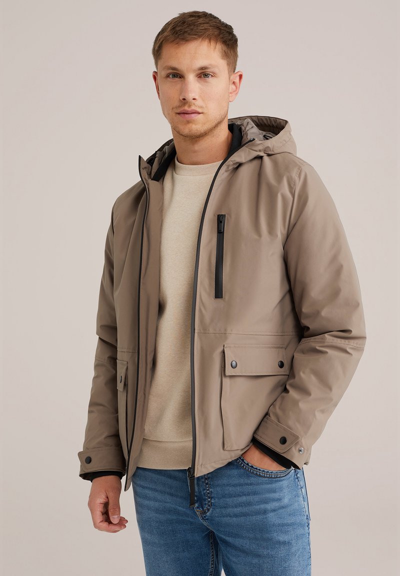 Chaqueta con capucha color arena de textura suave, con cierre frontal y bolsillos laterales. La lleva puesta sobre un suéter beige claro y lo combina con unos jeans azules.