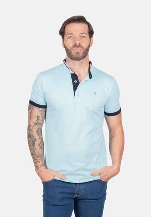 Hombre con barba y tatuajes que lleva un polo azul claro con ribete oscuro, de pie con las manos en los bolsillos frente a un fondo liso.