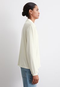 Lysegul langermet topp med krage med knapper. Har en avslappet passform og ribbestrikkede mansjetter, kombinert med blå denimjeans.