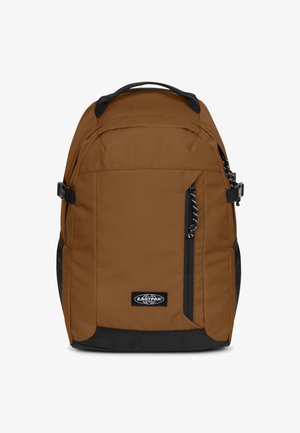 Mochila de tela marrón con detalles negros, cremalleras y un bolsillo delantero. Cuenta con correas ajustables y un asa superior para llevar.